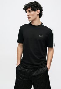 Fekete rövid ujjú póló kerek nyakkivágással, melyen a mellkason finom EA7 logó és a vállon szürke díszítés található.