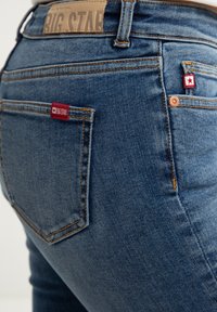 Sinine teksad, lähedalt pildistatud tagataskust. Omab pruunist nahast siltu ja punast silti logoga. Heledat tikandit ja tekstureeritud kangast.