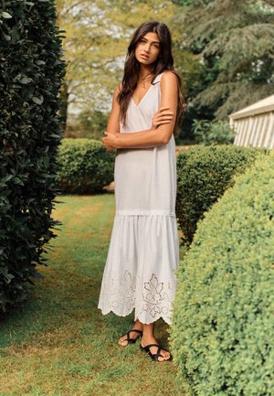 Donna in piedi all'aperto tra cespugli potati, indossa un vestito bianco senza maniche con orlo in pizzo e sandali neri, le braccia incrociate.