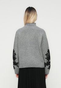 Femme avec des cheveux blonds raides portant un pull gris à col roulé avec de la dentelle florale noire sur les manches et une jupe plissée noire.