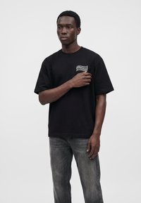 Homme debout portant un t-shirt noir à motif graphique et un jean gris délavé sur un fond clair uni.