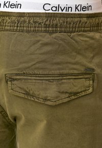 Maslinasto zelene pamučne hlače s bijelim elastičnim pojasom na kojem je oznaka "Calvin Klein" i jednim stražnjim džepom s preklopom.