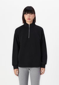Svart sweatshirt med glidelås og høy krage, ribbede mansjetter og avslappet passform. Stoffet ser glatt ut med en matt finish. Brukt med grå bukser.