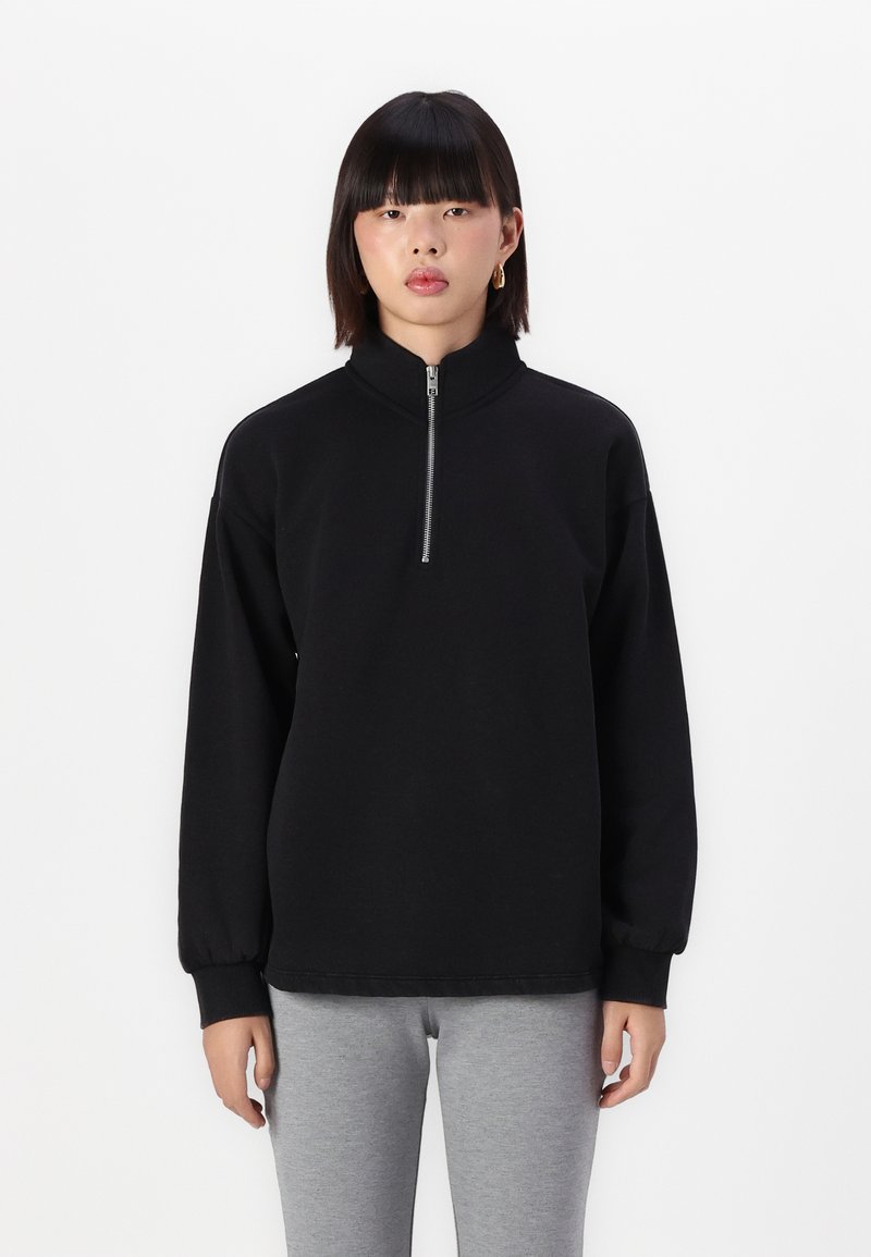 Svart sweatshirt med glidelås og høy krage, ribbede mansjetter og avslappet passform. Stoffet ser glatt ut med en matt finish. Brukt med grå bukser.