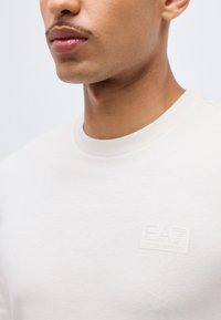 Νεαρός άνδρας που φοράει ένα απλό λευκό t-shirt EA7 Emporio Armani, το οποίο φαίνεται από το κάτω μέρος του προσώπου, τον λαιμό και το πάνω μέρος του στήθους με το λογότυπο στην αριστερή πλευρά.
