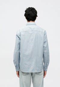 Ανοιχτό μπλε πουκάμισο με μακριά μανίκια και κουμπιά, με διακριτικό κάθετο ριγέ μοτίβο και καμπυλωτό τελείωμα. Το ύφασμα φαίνεται λείο και απαλό.