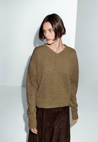 Donna con capelli corti castani, che indossa un maglione verde oliva a V largo e una gonna marrone scuro, in piedi davanti a uno sfondo bianco e grigio.
