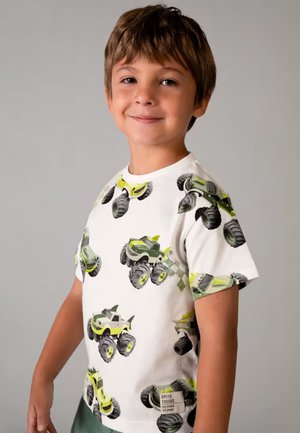 Ragazzo sorridente che indossa una maglietta bianca con stampe di monster truck verdi e grigie su uno sfondo grigio uniforme.