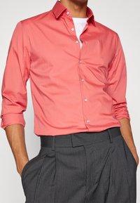Coral button-up shirt with long sleeves, subtle sheen, and white logo detail at chest, paired with dark pinstriped trousers.