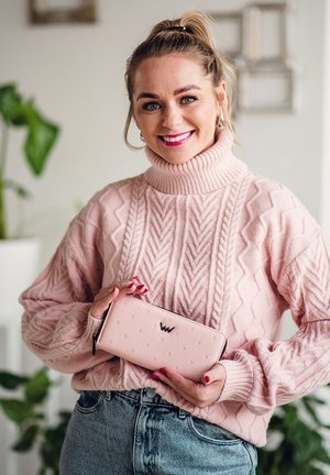 Woman in a pink cable-knit sweater holding a matching pink wallet, smiling, with plants and wall decor blurred in the background.