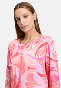 Blusa floreale con scollatura rotonda, maniche lunghe e una palette di colori rosa con motivi astratti in rosso e lilla, realizzata in tessuto leggero e morbido.