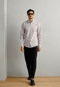 Λευκό πουκάμισο με μακριά μανίκια και λουλουδένιο μοτίβο, συνδυασμένο με μαύρο παντελόνι και μαύρα παπούτσια. Μοντέλο φορώντας γυαλιά ηλίου, που στέκεται σε ξύλινο πάτωμα.