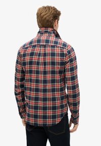 Camisa de cuadros en azul marino y rojo de manga larga con cuello abotonado, con dobladillo redondeado y puños, hecha de tela suave, con detalles de costura visibles.
