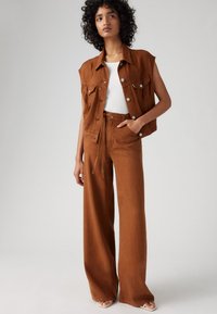 Ruskea denimpitkäliivi, jossa on etutaskut, yhdistetty korkeavyötäröisiin, leveälahkeisiin ruskeisiin housuihin. Mallilla on päällä valkoinen tiukka paita.