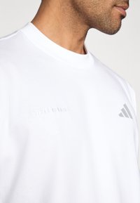 T-shirt branca de manga curta com texto em relevo "ADIDAS RUNNING", decote redondo e logótipo das três riscas características em cinza claro. Textura suave.