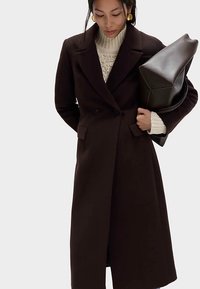 Brązowy płaszcz z mieszanki wełny o dwurzędowym kroju, z ostrymi klapami i bocznymi kieszeniami. W połączeniu z kremowym swetrem w prążki i dużą torbą.