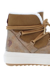 Hnědé semišové kotníkové boty s bílým umělým kožichem, béžovými tkaničkami, zlatým kováním a embosovaným logo detailem na straně. Texturovaný povrch a gumová podrážka.