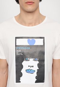 Weißes Baumwoll-T-Shirt mit einem grafischen Aufdruck eines Gesichts mit blauen Augen und blauen Lippen, umgeben von einem schwarzen Hintergrund und blauem Text.