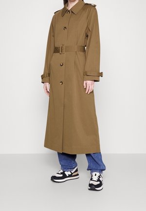 Persoon die een lange bruine trenchcoat met ceintuur draagt, blauwe spijkerbroek en zwart-witte sneakers, staand tegen een effen witte achtergrond.