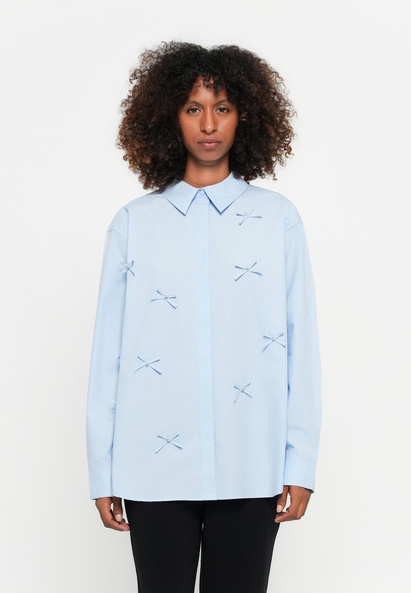 Camisa azul clara com colarinho pontudo, mangas longas e fecho frontal, apresentando laços decorativos com contas como acentos.