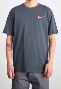 Szara bawełniana koszulka z małym wzorem czerwonej róży i zielonych liści, z białym haftowanym logo Nike po lewej stronie klatki piersiowej.