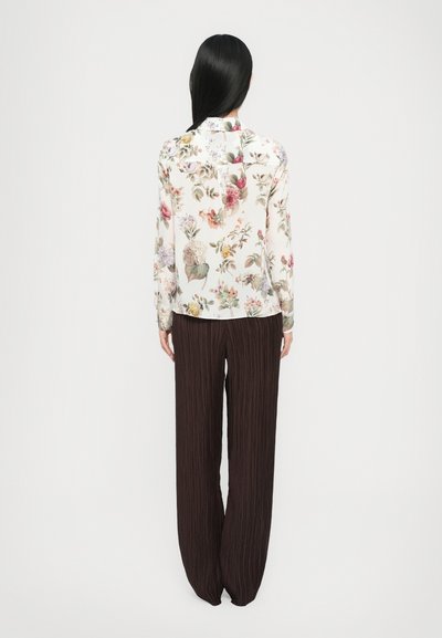 Bluse mit langen Ärmeln in Creme, verziert mit mehrfarbigen Blumenmustern. Kombiniert mit braunen, weiten plissierten Hosen.
