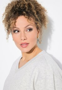 Sudadera de cuello en V gris claro con una textura suave, con hombros ligeramente caídos y un ajuste relajado, llevada por un modelo con cabello rizado.