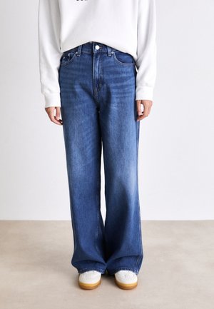 Jeans de mezclilla de pierna ancha en azul medio con un aspecto desgastado, con un cierre clásico de botón y cremallera, combinados con zapatillas blancas.