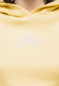 Personne portant un sweat à capuche jaune clair avec un logo "GAP" blanc centré sur la poitrine.