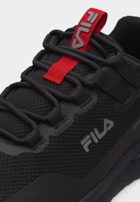 Baskets FILA noires en tissu texturé, logo gris sur le côté, lacets noirs et languette rouge avec le logo FILA sur la langue.