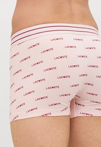 Lichtroze boxershorts met herhaalde rode "LAC♥STE" logo-print en een zachte elastische tailleband met rode en witte strepen.