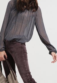 Průhledná modrošedá blůza s knoflíkovou légou a dlouhými rukávy, kombinovaná s švestkovými skinny džínami. Modelka drží béžovou kabelku.