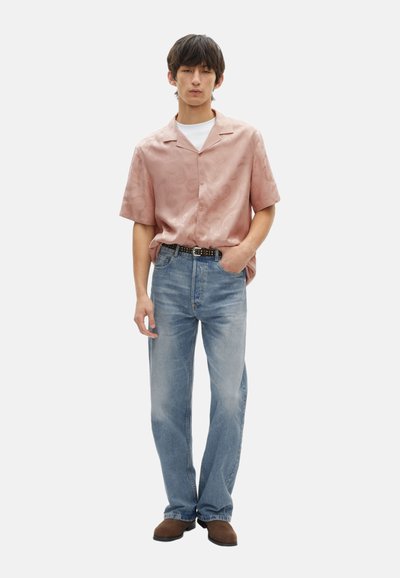 Korte mouwen, roze satijnen shirt met subtiel patroon, gedragen met lichtblauwe spijkerbroek met rechte pijpen en bruine schoenen.