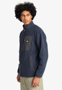 Giacca in pile blu navy con colletto alto, dotata di tasca sul petto con zip grigia e tasche laterali, tessuto testurizzato e chiusura con zip.