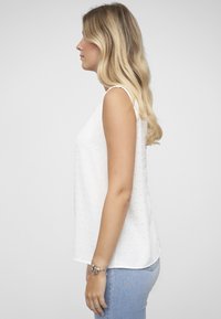 Witte mouwloze top met gestructureerde stippen, losse pasvorm, licht gebogen zoom, subtiele glans, gecombineerd met lichtblauwe spijkerbroek. Zijaanzicht.