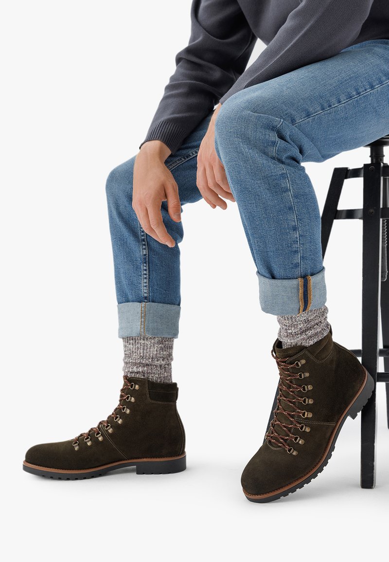 Botine din suede maro la glezna, cu ochiuri din metal, sireturi și talpă din cauciuc. Asortate cu blugi de culoare albastru deschis și șosete tricotate gri.