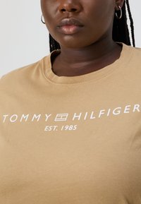 Béžové bavlnené tričko s okrúhlym výstrihom. Na prednej strane sa nachádza biele logo "TOMMY HILFIGER" a nápis "EST. 1985". Klasický strih.