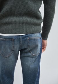 Maglione verde a coste indossato sopra una camicia bianca, abbinato a jeans di denim blu con cinque tasche e cuciture visibili sul retro.