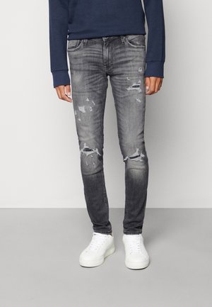 Homme portant un jean slim noir délavé et déchiré, des baskets blanches et une chemise bleu foncé à manches longues, debout sur un sol gris devant un mur clair.