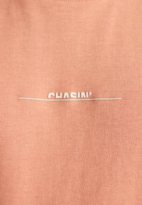 T-shirt leggero di colore pesca realizzato in tessuto morbido, con la scritta "CHASIN'" in lettere bianche e un sottile dettaglio a linea orizzontale.
