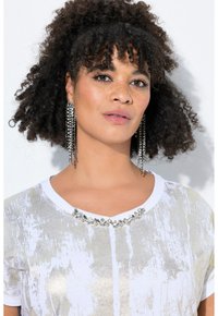 T-shirt bianco con motivi verticali dorati simili a vernice e un colletto incastonato di gioielli. Il modello indossa orecchini lunghi e scintillanti; capelli ricci e texturizzati.