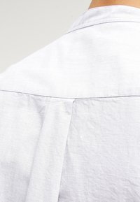 Camisa de botón gris claro con un diseño de espalda plisada, hecha de un tejido texturizado, que presenta un cuello clásico y un dobladillo recto.