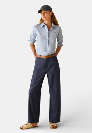 Donna che indossa una camicia azzurro chiaro abbottonata, pantaloni larghi scuri con cintura marrone, sandali beige con punta aperta e un cappellino da baseball scuro, in piedi in posizione rilassata.