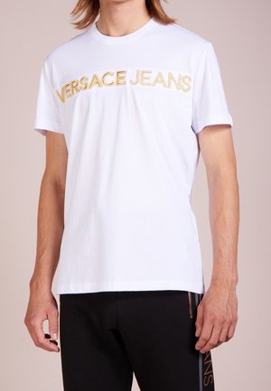 Personne portant un t-shirt blanc Versace Jeans avec lettrage doré et un pantalon noir avec une bande verticale dorée sur le côté.