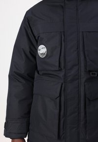 Svart vadderad jacka med en slät yta, flera framfickor och en rund logotyp-patch på vänster bröst. Designen inkluderar högt krage och justerbara ärmslut.