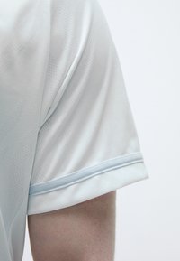 Gai zila sporta krekls ar gludu tekstūru, ar izteiksmīgu rakstu un kontrastējošu gaiši zilu apdari gar piedurkņu malām.