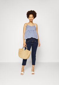 Top sem mangas azul e branca às riscas com bordas franzidas, jeans escuros e uma bolsa bege tecida com alças de contas. Sapatos de abertura na frente creme.
