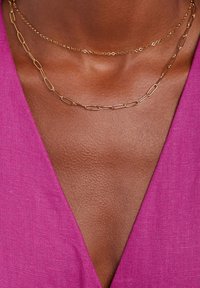 Gouden gelaagde kettingen met ovale schakels en een delicate ketting, voor een getextureerde huidtoon, onder een diep V-hals roze kledingstuk.