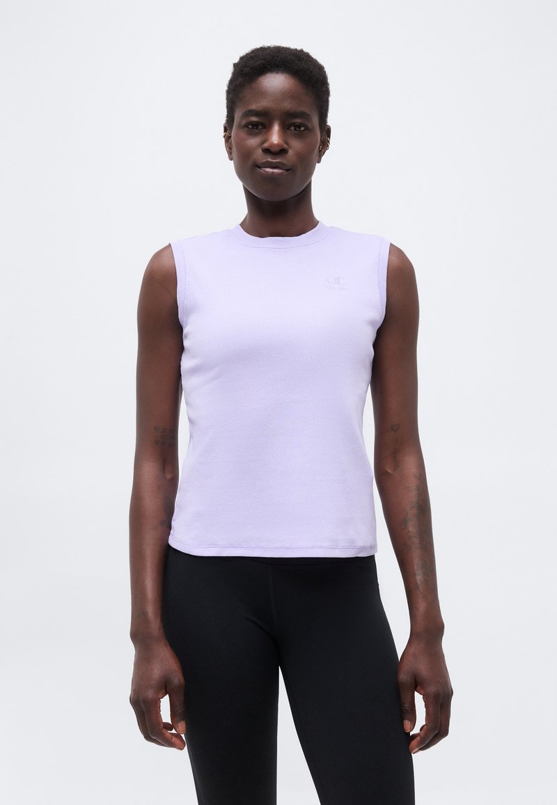 Γυναίκα που στέκεται μπροστά και φοράει ένα αμάνικο ανοιχτό μωβ τοπ και μαύρα κολάν, με φόντο ένα απλό λευκό τοίχο.