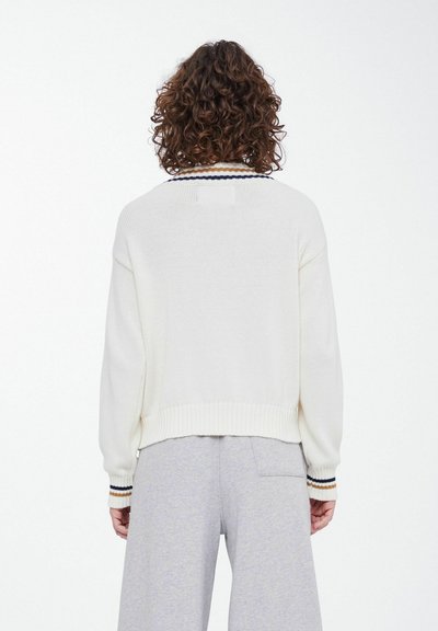 Cream sweater with ribbed cuffs featuring navy, white, and tan stripes. Loose fit with a solid texture, worn over grey sweatpants.